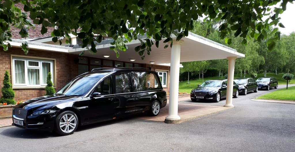 A polished black hearse waiting beneath a crematorium portico, with family limousines lined up behind it
