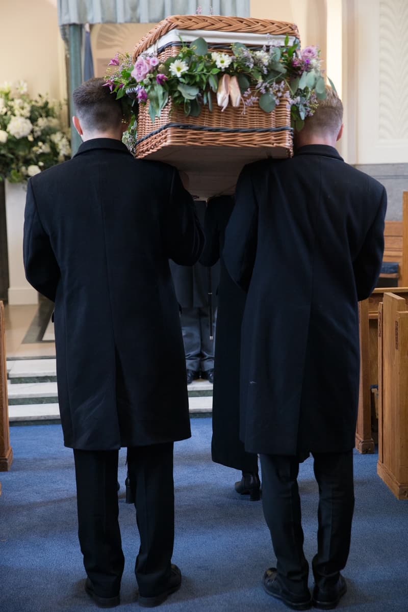 Two bearers carrying a woven wicker coffin, dressed with fresh flowers, into a chapel